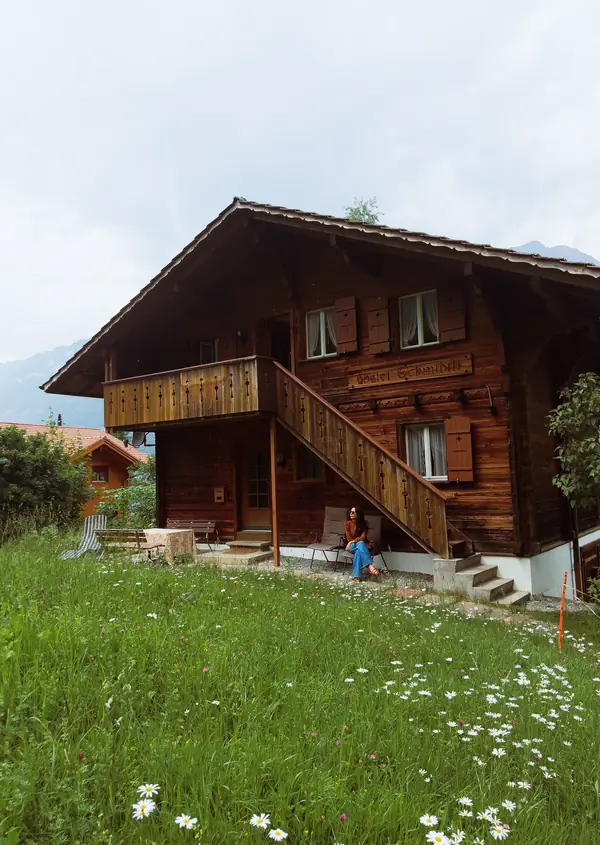 A dark timber chalet with a long balcony, on a meadow of daisies below the hills