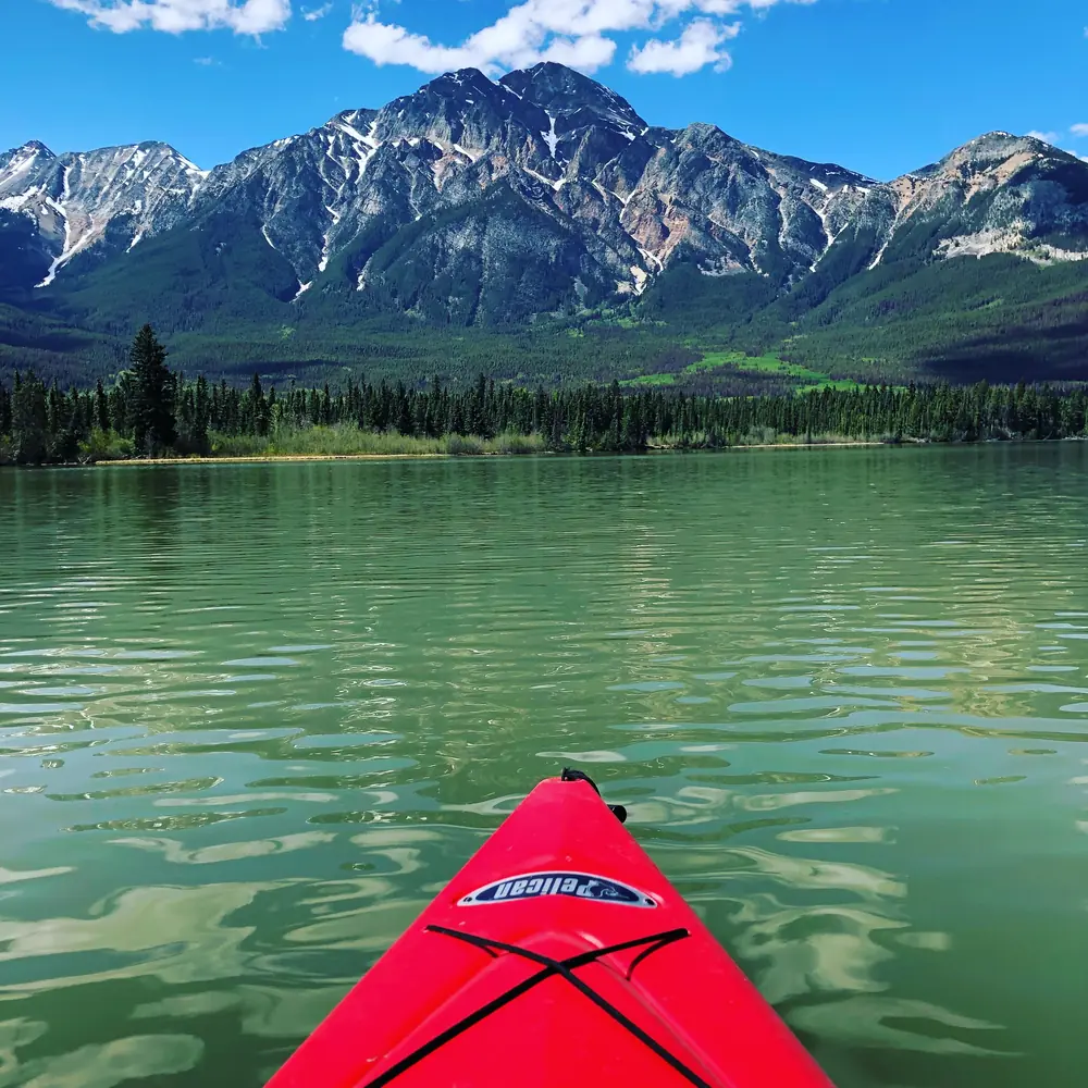 The red bow of a kayak on a clear green lake, jagged snow-streaked mountains ahead