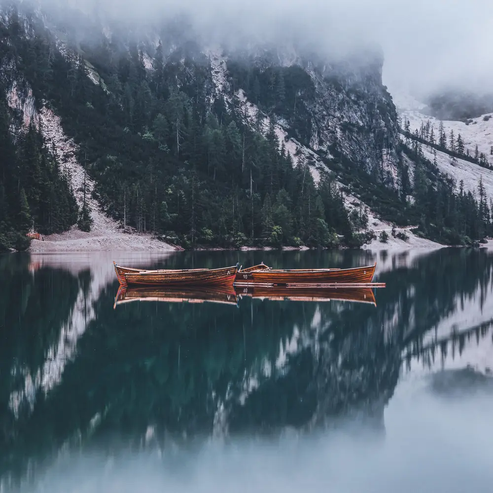 Two rowing boats on a still lake in the mist, the forested mountainside mirrored in the water