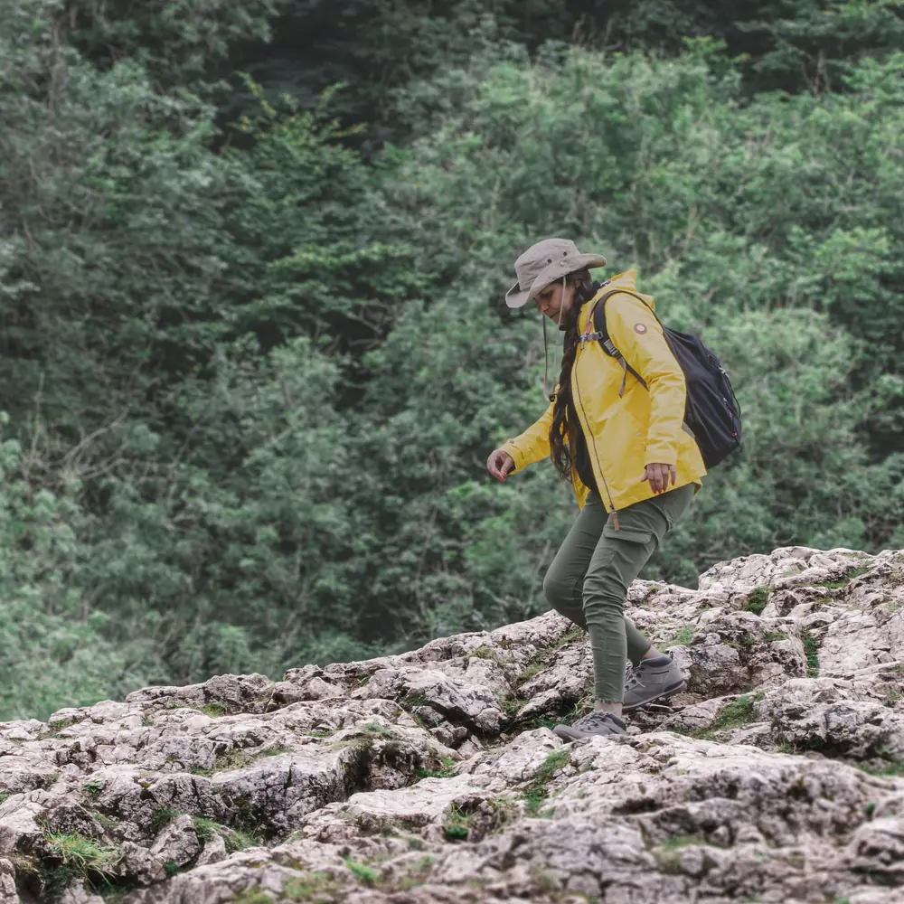 A walker in a yellow jacket and a sun hat crossing bare rock with a rucksack, pines behind