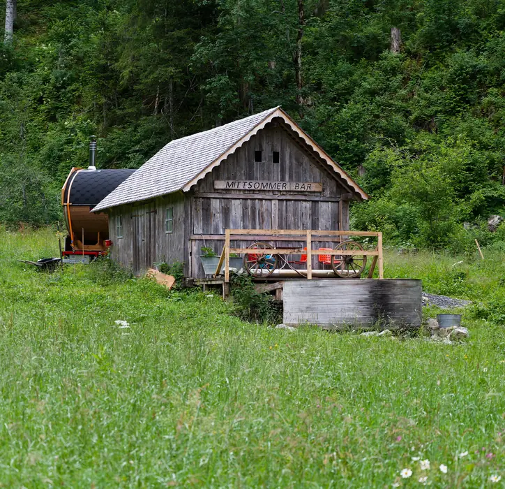 A barrel sauna and a wooden hot tub beside an old timber hut in a green meadow below the forest