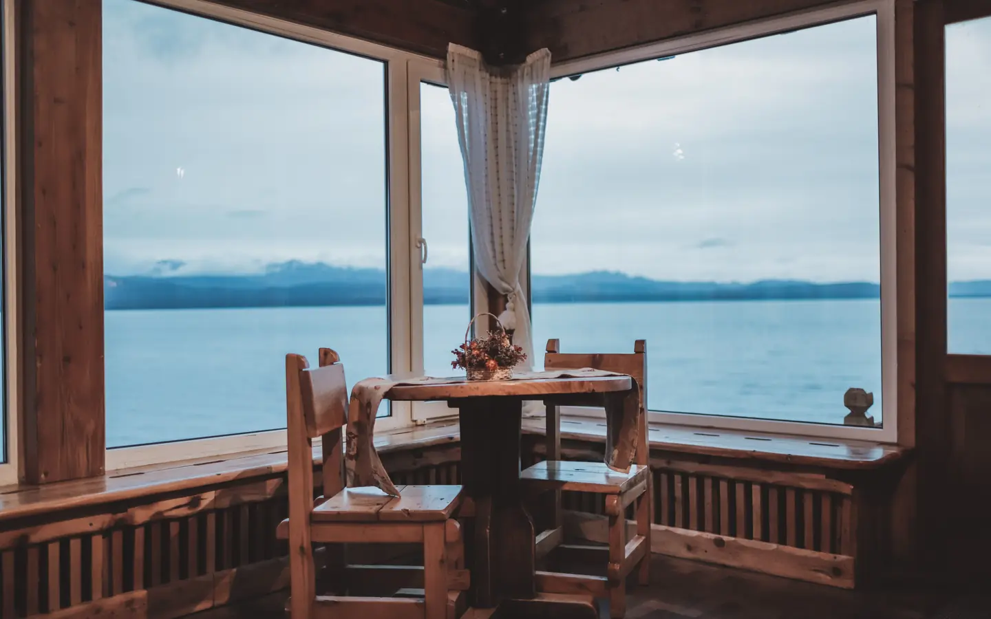 A small table and two wooden chairs in a corner window over wide, still water and distant hills