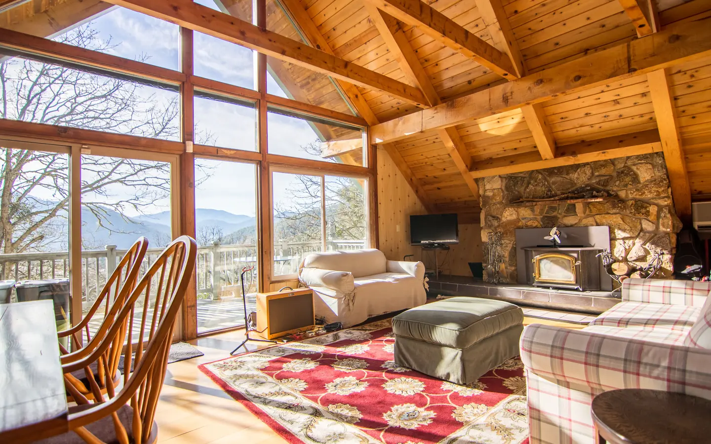 A timber-framed sitting room with a stone fireplace, armchairs, a red rug and tall windows onto the hills