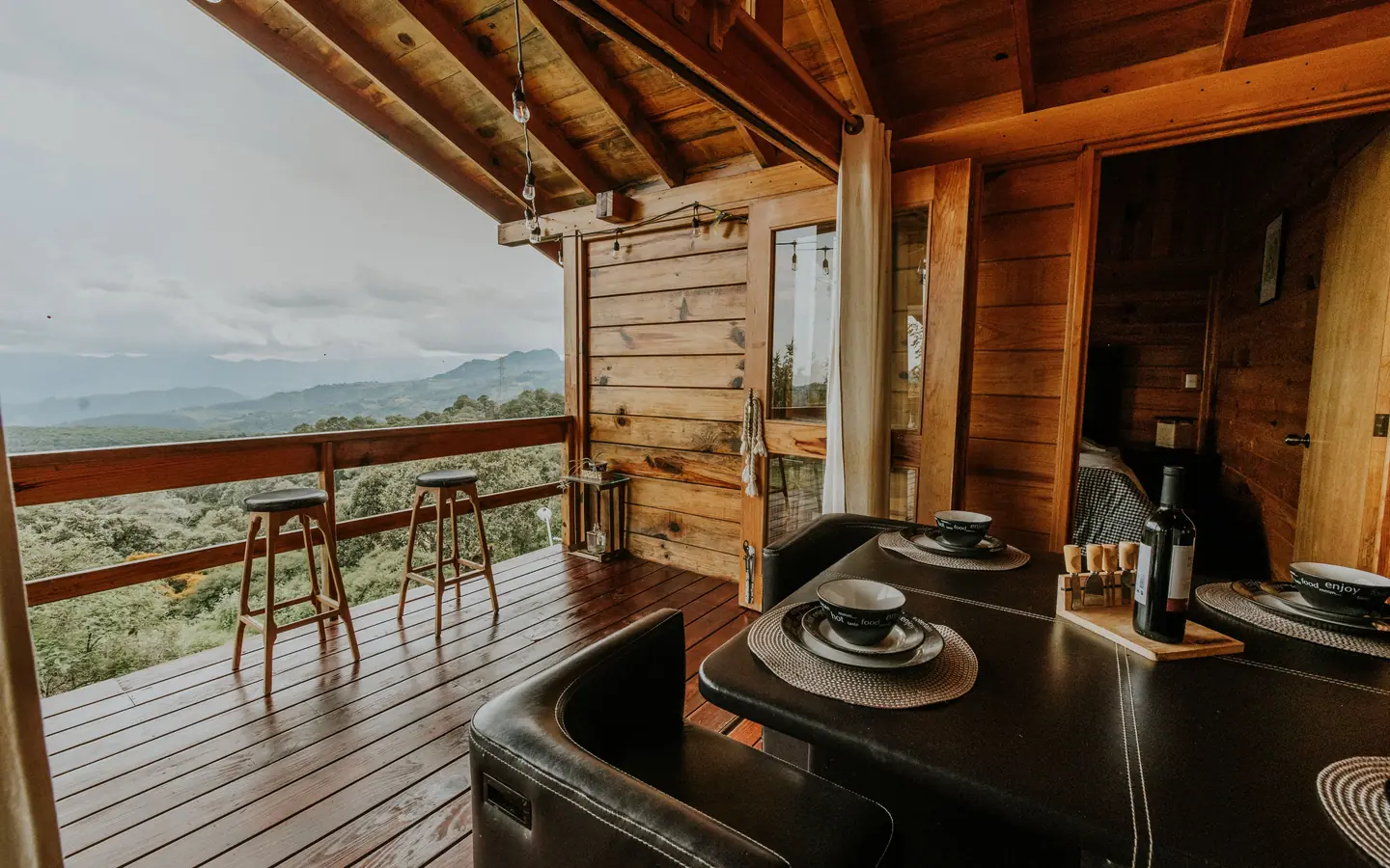 A wooden deck with high stools and a dining table under a timber roof, looking over forested hills