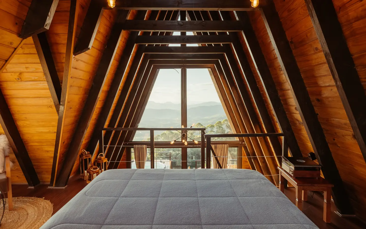 A wide bed facing the glass end wall of an A-frame cabin, hills beyond the balcony